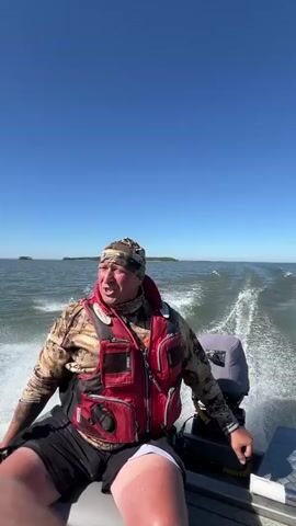 A man in a red life vest and camouflage clothing is sitting in a boat, holding onto the steering wheel. The boat is moving at a high speed across a body of water, creating a wake behind it. The sky is clear and blue, and there are islands in the distance. The man appears to be speaking, but his words are obscured by the sound of the engine.