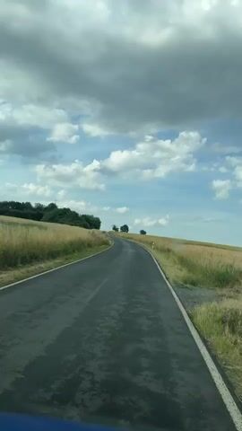 A first-person perspective video shows a road curving through a rural landscape with fields of dry grass on either side. The sky is partly cloudy with patches of blue. The road appears to be asphalt with a white line marking the edge. There are occasional trees and bushes visible along the sides of the road and on a distant hill. The video has a slightly muted color tone, suggesting it may have been filmed on an overcast day. No people or other vehicles are visible in the frame. The camera is positioned as if it is inside a vehicle, looking forward.