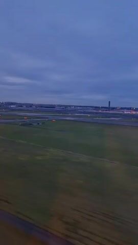 The video shows an airplane landing at an airport on a cloudy day. The camera is likely inside the airplane, looking out of a window. We can see the runway and taxiways, with lights marking the edges. There are other airplanes in the distance. The ground is mostly grass.