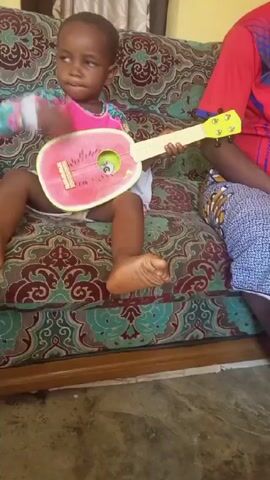 A young child sits on a patterned sofa strumming a watermelon-shaped toy guitar. The child has short, dark hair and is wearing a long-sleeved shirt with a colorful floral design and white shorts. An adult wearing a red shirt and patterned pants sits next to the child on the sofa. The sofa has a brown wooden frame and patterned upholstery featuring shades of green, brown, and white. The floor beneath the sofa is a light, possibly concrete or tiled surface. A portion of a window and curtain are visible in the background. The child looks at the camera with a serious expression while playing the toy guitar.