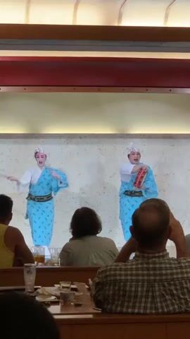 The video shows a performance on a raised platform with a beige background. Two performers, dressed in light blue traditional Japanese robes with a white trim and white hats, are on the platform. The performer on the left is holding two thin sticks. The performer on the right is holding a red and white object and a drum. The performers are engaged in a coordinated movement, possibly a dance or a martial arts demonstration. In the foreground, several audience members are seated at tables, facing the performance. They appear to be in a restaurant setting, with glasses and dishes on the tables. The lighting suggests an indoor environment.