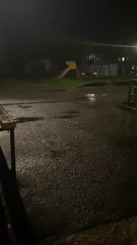 The video shows a nighttime outdoor scene with a playground in the background. The ground is wet, indicating recent rain, with puddles reflecting the light from a nearby streetlamp. A metal gate and some foliage are visible on the right side of the frame. The playground equipment, including a slide, is dark but illuminated by the ambient light. A bench is also present. The sky above the trees shows a faint purple glow, possibly from distant lightning. The camera appears to be positioned at ground level, looking slightly upwards and across the scene.