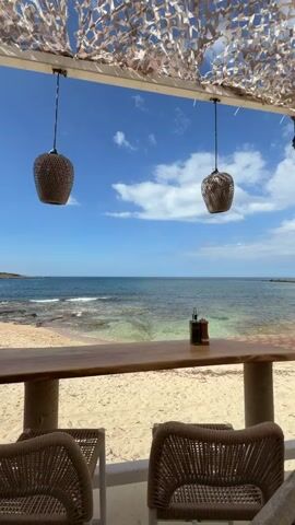 A static, eye-level shot shows a beachside bar with a stunning view of the ocean. The bar is constructed from light-colored wood with woven chairs set up in front of it. On the bar, there are condiments. Above, a patterned canopy offers shade while allowing glimpses of the blue sky dotted with white clouds. Two decorative, woven light fixtures hang from the canopy. The sandy beach stretches to the calm, turquoise water. In the distance, a small rocky island is visible.