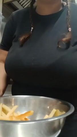 A woman tosses seasoned french fries in a metal bowl. The woman is wearing a black shirt and has braids. The kitchen background shows stainless steel surfaces.