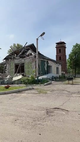 The video shows a building that has been heavily damaged. The roof is collapsed, and there are piles of debris. A tall brick tower stands to the side of the building. The building appears to be located in a residential area, as there is grass and a street in front of it. The sky is blue with some clouds. There is a person speaking in the background, but they are not visible in the frame.