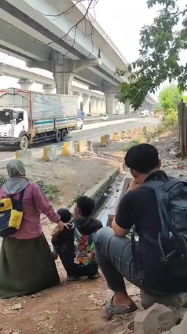 The video shows a street scene under a large overpass. In the foreground, a woman wearing a hijab and a pink jacket is holding the hand of a child who is being carried by another person who is facing away from the camera. To the right, another person is sitting down facing away from the camera. In the background, there is a large truck with a white cargo box, followed by a white SUV and another truck with a blue tarp covering its cargo. The area around the people in the foreground appears to be a dirt path with some debris. The overpass has multiple lanes of traffic.