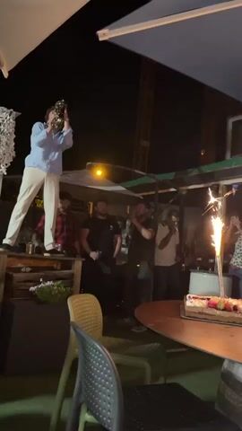 The video is a nighttime outdoor scene. A young man in a light blue collared shirt and cream-colored pants stands on a raised wooden platform, playing a saxophone. He is looking down towards the camera. Several other men are standing behind him and to his right, appearing to watch him or the scene. To the right of the frame, a cake with lit sparklers is visible on a table. The environment appears to be a gathering or party, possibly a restaurant or bar patio, with umbrellas and lights suggesting an evening event. The overall mood is festive.