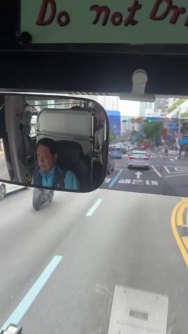 The video shows a view from inside a vehicle, likely a bus or large van, looking forward through the windshield and into a side mirror. In the side mirror, a middle-aged East Asian man with dark hair is visible. He is wearing a light blue vest over a darker shirt and appears to be the driver. He looks towards the camera. The windshield shows a street scene with other vehicles, buildings, and traffic signals. The road has lane markings, including an arrow pointing forward and some Korean text. The overhead interior of the vehicle has a green sign with red lettering that reads "Do not Dri".
