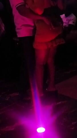 The video shows a group of people dancing in a dimly lit nightclub, illuminated by colorful disco lights. The main focus is on a woman wearing a short, patterned dress with ruffles, dancing closely with a man wearing a white shirt. The camera angle is low, emphasizing the movement of the dancers. Other individuals are partially visible in the periphery, including one in a plaid shirt. The atmosphere is lively and energetic, typical of a nightclub setting.