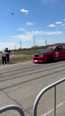 The video shows a red car performing a burnout on an asphalt track. Two individuals stand on the side, seemingly observing the car. Power lines and some greenery are visible in the background under a bright sky with scattered clouds. The car produces a large cloud of smoke as it spins its wheels.