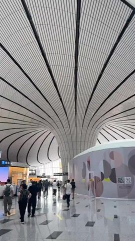 The video shows the interior of a modern airport. The ceiling is a prominent feature, with a white, ribbed design that curves and swoops, creating a dramatic effect. Natural light filters in through a series of skylights integrated into the ceiling. The floor is polished and reflective, showing the reflections of the architecture and the people.

Several people are visible walking through the spacious terminal. They are dressed in casual clothing. Some are carrying luggage, suggesting they are travelers. There are shops and information kiosks visible in the background. One shop appears to be a Louis Vuitton store, indicated by signage. There are also digital screens and information boards throughout the terminal.

The overall impression is of a large, bright, and architecturally striking public space, likely an airport terminal, bustling with travelers.