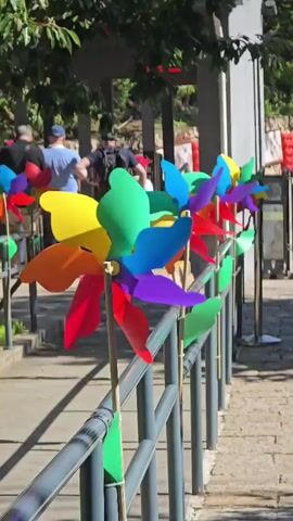The video shows a close-up of several colorful pinwheels attached to a metal railing. The pinwheels are in various stages of rotation, suggesting a gentle breeze. In the background, several people are walking up a sloped path lined with more pinwheels. The environment appears to be outdoors with trees and foliage visible. The lighting suggests it is daytime.