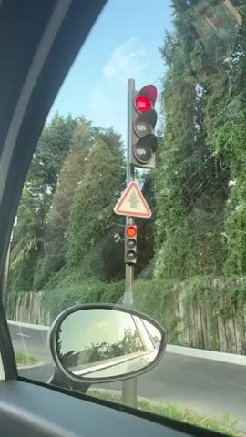 The video is filmed from the interior of a white Fiat 500, looking out the front windshield and side window. The car appears to be stopped at a traffic light, as evidenced by the red light visible in the first frame. The traffic light is on a pole, accompanied by a triangular road sign. Beyond the traffic light, a road stretches ahead, lined with trees and buildings. The sun is low in the sky, suggesting it is either early morning or late afternoon. The scene is calm and appears to be in a suburban or urban area.