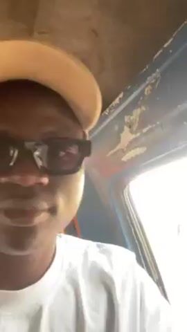 A young man with dark skin, wearing a white t-shirt, a light brown baseball cap, and black sunglasses, is recording a video of himself inside a vehicle. He glances at the camera and then makes a peace sign with his fingers. The interior of the vehicle appears old, with a blue dashboard and worn paint on the ceiling. The background suggests an outdoor setting, with light streaming in through the window. There is ambient sound, possibly street noise.