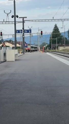 A train approaches a station platform. The train is silver and red and has multiple cars. The platform is paved with asphalt and has white lines marking the edge. There are benches and trash receptacles on the platform. A person is visible sitting on a bench, facing away from the camera. Overhead, there are train signals and electrical wires. In the background, there are buildings and trees, and mountains are visible in the distance. The sky is cloudy. A red and black scooter is parked on the left side of the platform. A blue bag is hanging from a pole on the left side of the platform.