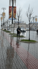 City worker trims grass around tree during rainy morning