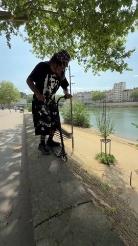 A person with dreadlocks, wearing a bucket hat, black t-shirt, and patterned shorts, performs a stunt on a kick scooter. The video begins with the individual positioned on a concrete wall or ledge, preparing to ride the scooter down a steep, rough-textured slope. In the background, a wide river, a dirt path, and urban buildings are visible under a bright, clear sky. The person then pushes off, successfully riding the scooter down the slope and landing on the path below. Another person stands on the path, observing the action. The scooter rider continues along the path, moving away from the camera. The scene depicts an urban riverside environment, likely in Lyon, France. No visible text is present.