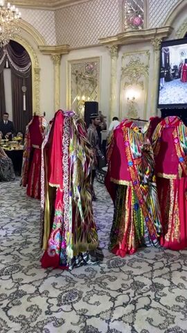 A group of dancers wearing brightly colored, ornate traditional clothing are performing in a ballroom. The dancers are mostly women, with their faces obscured by veils or elaborate headdresses. They are moving in a synchronized manner, with their arms extended. The ballroom is richly decorated with gold accents, chandeliers, and patterned carpets. In the background, guests are seated at tables, observing the performance. A large screen is mounted on the wall, displaying an image. The lighting is dim, creating a festive atmosphere.