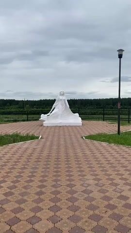 The video shows a white statue of a woman from the waist up, appearing to be wearing a flowing gown or cape. She is standing in front of a dark, dense forest under a cloudy sky. The statue is centered on a paved area with a distinctive diamond-shaped pattern. The paved area leads to a pathway with the same pattern, which narrows as it approaches the camera. To the right of the statue and pathway, a black metal fence encloses the area. A single black lamppost is visible to the right of the statue, near the fence. The ground around the paved area is covered in grass. The video is filmed from a low angle, looking up at the statue.