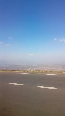 The video shows a view from a moving vehicle on a highway. The sky is blue with a few clouds. In the distance, a hazy view of a town or agricultural area can be seen. A silver car passes by.  A small roadside shack with two people standing near it comes into view. The road has white lane markings.