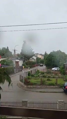 The video opens with a slightly blurred view of a residential street during what appears to be rainy weather. The camera is positioned from inside a building, looking out through a window that has water droplets on it. In the foreground, there is a dark railing. Beyond the railing, the street is visible, wet from rain, with a few cars parked and driving. Houses with gardens are lined along the street, surrounded by trees and fences. One person can be seen walking on the sidewalk. The sky is overcast and a dense fog or cloud obscures the upper part of a large tree in the distance. The overall atmosphere is one of a quiet, drizzly day in a suburban neighborhood.
