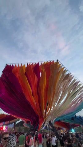 The video shows a large crowd of people at what appears to be an outdoor festival or concert. Overhead is a massive structure adorned with colorful fabric strips that create a tent-like effect. The strips are predominantly in shades of red, orange, and yellow. The sky above is a pale blue with scattered clouds. The camera angle is low, looking up at the overhead structure and capturing the energy of the crowd below.  Occasional flashes of pink light occur.
