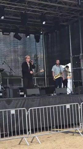 A band performs on an outdoor stage. A male singer in a black jacket and glasses stands center stage, holding a microphone. To his right, a man plays a saxophone. In the background, a drummer is visible behind a drum kit. The stage is set up with musical equipment, including speakers and microphones. A black curtain is behind the band, and a metal barricade is in front of the stage. The overall environment appears to be a daytime outdoor event.