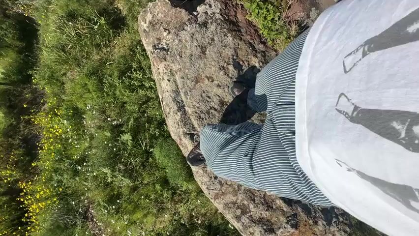 The video begins with a low-angle shot focusing on the lower body of a person standing on a rugged, reddish-brown rock. The person is wearing striped pants, possibly dark blue or green with white stripes, and dark shoes. To the left of the rock, a vibrant green grassy area is visible, adorned with numerous small yellow wildflowers.

As the video progresses, the camera slowly pans upwards and outwards, revealing a expansive coastal vista. The deep blue sea stretches across the horizon under a clear, bright blue sky. In the distance, a prominent dark landmass, resembling a volcanic island, rises dramatically from the calm waters. The grassy cliff edge, where the person is positioned, remains visible on the right side of the frame. The scene suggests a sunny, clear day with excellent visibility. The person's white t-shirt, featuring a faint graphic design, is also partially visible. The setting appears to be a scenic lookout point, possibly on Lipari, Italy, offering views of the sea and distant islands.