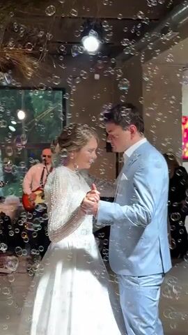 A wedding couple dances while surrounded by bubbles. The bride wears a long-sleeved, beaded white wedding dress and her hair is pulled back. The groom wears a light blue suit. A band plays in the background. The environment appears to be an indoor venue with some greenery visible through windows.