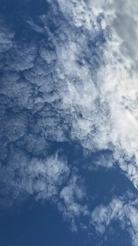 The video shows a low-angle shot of a cloudy sky over the ocean. The clouds are white and fluffy in some areas and more wispy and gray in others. The sun is partially obscured by clouds, creating a soft, diffused light. In the distance, two islands or peninsulas are visible on the horizon. The ocean surface is choppy, with small waves reflecting the light. The video appears to have been filmed from a boat.