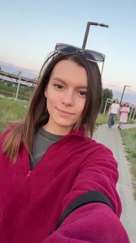 A young woman with brown hair and sunglasses perched on her head is taking a selfie outdoors. She is wearing a dark pink fleece jacket over a grey t-shirt. The background shows a grassy area with some trees and a fence. Two other people, one in a white shirt and another in a pink shirt, are walking away in the distance. The sky is a soft blue with hints of pink, suggesting it is either sunrise or sunset.