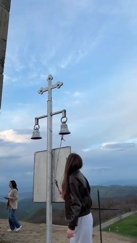 The video shows a young woman ringing two small bells hanging from a metal post with a cross on top. The post also has a white, blank sign attached. The woman is wearing a brown leather jacket and a long gray dress. Behind her is a vast landscape of green hills under a blue sky with white clouds. To the left, a woman in a tan jacket and jeans walks by. The camera then pans up to show a large, gray concrete cross made of stacked blocks, with a small shrine attached to its base. A man with a black jacket and a small bag is seen taking a photo with his phone.