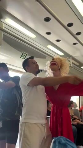 A man in a white t-shirt and light-colored pants stands on a train, holding a puppet with blonde curly hair and a red dress. He is moving the puppet's arms and head. The puppet's mouth is open, and its eyes are wide. The man is smiling and looking at the puppet. There are other people on the train sitting in the background. The train appears to be in motion.