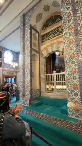 This media file captures an interior view of a mosque, likely in Eyüpsultan, Turkey, given the context. The scene is filled with ornate Islamic tilework featuring intricate floral and geometric patterns in shades of blue, red, and green. A plush, turquoise carpet covers the floor and rises in tiers, forming a raised platform.

In the foreground, a person wearing a dark hijab with a colorful stripe and a studded jacket is seen from behind, with their hands clasped in prayer. Other individuals, some also in headscarves, are visible in the background, suggesting a place of worship or pilgrimage.

Dominating the right side of the frame is a white marble tomb or cenotaph, adorned with golden calligraphy at its peak. This structure is likely the focal point of veneration within the mosque. To the left of the tomb, a man wearing a white skullcap is seated at a wooden desk, appearing to read or study. A microphone stands on a pedestal in front of him, hinting at a possible recitation or address.

Archways and wooden lattice screens add to the architectural richness of the space. Natural light streams in from windows, illuminating the scene, while ornate chandeliers also provide interior lighting. The overall atmosphere appears reverent and peaceful, typical of a religious site. Red velvet ropes and stanchions are present, demarcating visitor areas. Visible text on a red ribbon reads "KÜLTÜR VE TURİZM BAKANLIĞI," which translates to "Ministry of Culture and Tourism" in Turkish, further confirming the location as a significant cultural and tourist site in Turkey.