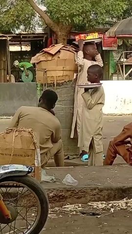 The video shows a street scene with several individuals. One young boy, dressed in a light-colored robe, is holding a plate of food at neck level. Another boy stands behind him with what appears to be a box on his head. A man in a light-brown outfit sits on a curb next to a motorcycle and a cardboard box. Another man in a brown outfit sits on the other side of the curb. Another man in a yellow soccer jersey stands talking to the boy with the plate, and another man stands to the side. Several buildings and trees are visible in the background. The audio includes traffic sounds.