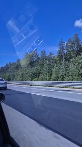 A white Audi sedan drives on a highway. The view is from another car, looking out the passenger side window. Tall green trees line the side of the highway, and the sky is a clear, bright blue with a few scattered clouds. There is a white stripe marking the edge of the highway. A metal overhead gantry is visible in the distance. The reflection of a smartphone is visible in the window.