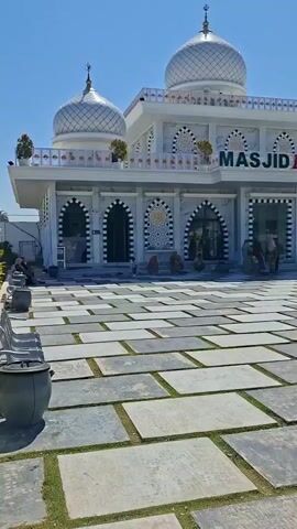 The video shows the exterior of a white mosque with silver domes, labeled "MASJID HARTON." The mosque has arched entrances and decorative patterns. In the foreground, there is a paved courtyard with large gray square tiles interspersed with grass. Several gray benches and trash receptacles are visible in the courtyard. A few people are seated near the mosque entrance and walking in the courtyard. The sky is clear and blue. The environment appears to be an open outdoor space.