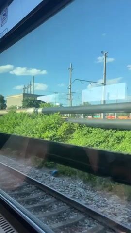 A view from a train window shows a bright blue sky with scattered clouds. The train tracks are visible in the foreground, with gravel ballast. In the middle ground, there are large industrial pipes, some black and some weathered, running horizontally. These pipes are partially obscured by lush green vegetation. Tall, grey utility poles with overhead electrical wires stand vertically in the background. The scene appears to be an outdoor industrial or transportation area on a sunny day. Reflections are visible on the train window.