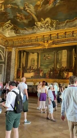 The video shows a wide shot of a grand hall with ornate decorations. The ceiling is a fresco depicting mythological scenes with cherubs and horses. A large, framed painting hangs on the wall, showing a banquet or feast scene within an architectural setting. The walls are adorned with marble and gold trim. Several people, dressed in casual summer clothing, are standing and walking around the hall, appearing to be tourists. Some are looking at the artwork, while others are conversing. The lighting suggests it is daytime. The floor is made of polished wood. The overall impression is of a museum or historical palace interior.