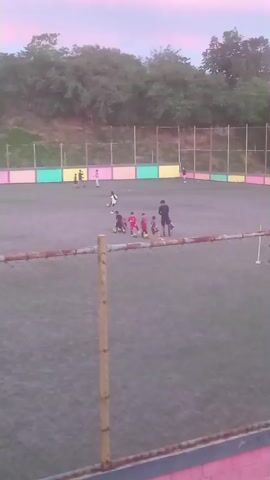 The video shows a group of people, likely children and adults, on a soccer field at dusk. The field has artificial turf and is enclosed by a fence. The sky is a pale pink and purple. There are several people scattered across the field, some appearing to be participating in a soccer practice. In the foreground, a chain-link fence with wooden posts is visible. In the background, there are trees, a hill, and buildings with colorful striped walls. The overall impression is an outdoor sports activity taking place as daylight fades.
