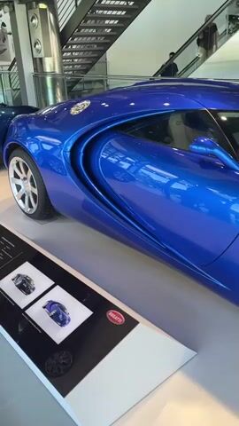A close-up shot of the side of a shiny blue Bugatti sports car. The car is on display indoors, with a staircase visible in the background. The car's sleek lines and distinctive design are highlighted. A sign with the Bugatti logo is visible at the bottom left. There are people in the background, but they are out of focus.