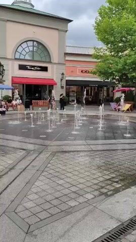 This is a video of an outdoor shopping area with a fountain in the foreground. The fountain has multiple jets of water spraying upwards. Behind the fountain are storefronts, including Tommy Hilfiger and Coach. Several people are visible in the video. One person is standing near the fountain, and others are walking or sitting. The sky is overcast. The video is filmed from a stationary perspective.