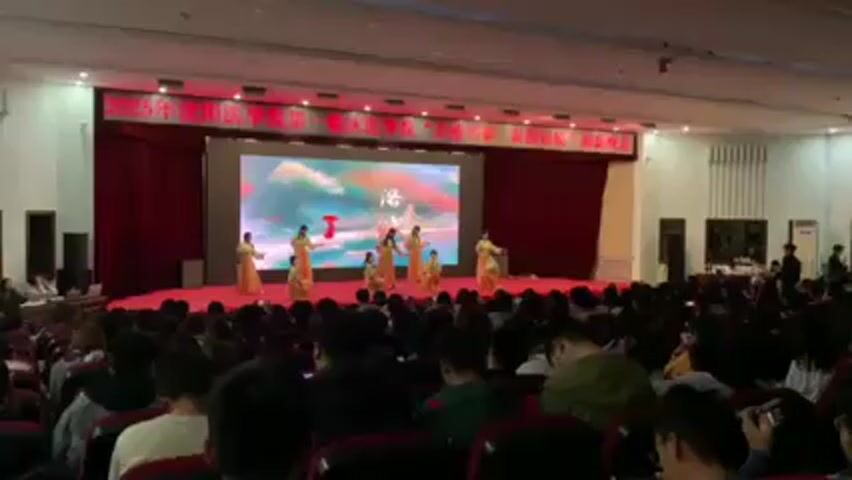A wide shot depicts a dance performance occurring on a stage in front of a seated audience. The stage is covered in red carpeting, and a large LED screen displaying animated artwork is positioned at the back. Red curtains frame the stage area. Seven performers, all women, are on stage wearing identical long-sleeved, gold-colored dresses. They are engaged in a choreographed dance routine. The auditorium is filled with people seated in red chairs, with their backs visible to the camera. The room is well-lit, and the stage area is brightly illuminated. The ceiling is white with recessed lighting. A banner in Chinese characters hangs above the stage, and tables with equipment and people are visible on either side of the stage. The overall atmosphere appears to be formal and celebratory.