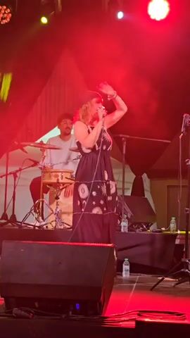 A medium shot captures a woman singing on a stage at night. She wears a black dress with a celestial pattern, a headscarf, and armfuls of bracelets. Her right arm is raised and extended, and her mouth is open as she sings into a microphone held in her left hand. Behind her, a man plays a drum set. The stage is lit with red and green stage lights. There are speakers and microphones visible on the stage. The background is dark and appears to be an outdoor venue with a wooden structure and some tents.