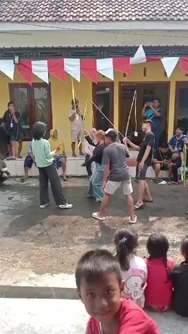 The video captures an outdoor event, possibly a celebration or competition, taking place in front of a yellow building adorned with red and white pennant banners. In the foreground, several children are seated, facing away from the camera, watching the activity. The central focus of the video is a group of adults engaged in what appears to be a game. One individual, holding a microphone, seems to be acting as an announcer or host. Several participants are holding long, thin poles with what look like small objects attached to the ends, possibly part of a game. One person is mid-stride, suggesting movement or action within the game. Other individuals are standing around observing the game. The ground is paved with a rough surface, and there are some scattered leaves. The overall atmosphere suggests a lively community gathering.