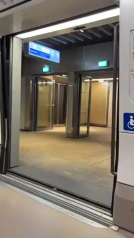 The camera shows a stationary view from inside a subway car, looking out at the platform through open doors. On the platform, there are multiple glass doors arranged in a row, with the light inside the train flickering from white to red. A blue sign points toward the exit. The platform is clean and modern. The doors of the subway car remain open during the entire video.