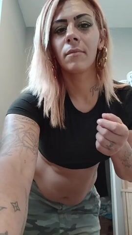 A woman stands in a bathroom, looking directly at the camera. She has fair skin, pink-tinged blonde hair, and green eyeshadow. She is wearing large gold earrings, a black crop top, and camouflage pants. Tattoos are visible on both arms and her stomach. She initially holds her fingers up, then adjusts her pants. The bathroom has gray walls, a white door, and a white shelf holding household items.  A sign reading "PLEASE HELP YOURSELF" is visible on the wall. The camera angle is slightly low, shooting up at the subject.