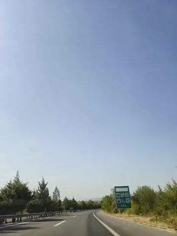 The video shows a view from a moving vehicle on a highway. The sky is clear and blue. On the right side of the road, there is a green sign with various icons indicating services such as restrooms, food, a petrol station, and disabled parking. The distance to these services is indicated as 8 km. The road is paved and has white lane markings. Trees and vegetation line both sides of the road. The vehicle is traveling at a moderate speed.