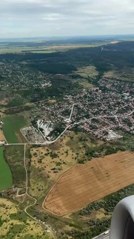 This is an aerial view from an airplane window of a rural landscape. In the foreground, the edge of an airplane engine is visible. The landscape below is a mix of green fields, brown agricultural fields, and a densely populated village with red-tiled roofs. A road winds through the village and surrounding fields. Further in the distance, the landscape becomes greener and more forested, with rolling hills extending to the horizon under a partly cloudy sky. There are no people visible in the frame.