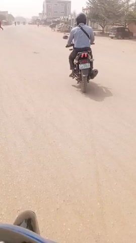 The video shows a person riding a motorcycle down a dusty, sandy road in what appears to be a town. The rider wears a helmet, a light-colored shirt, and dark pants, with a bag slung across their body. The motorcycle is black. Other vehicles, including other motorcycles, cars, and a tuk-tuk, are visible in the distance. Buildings line the road. The sky is hazy and bright.